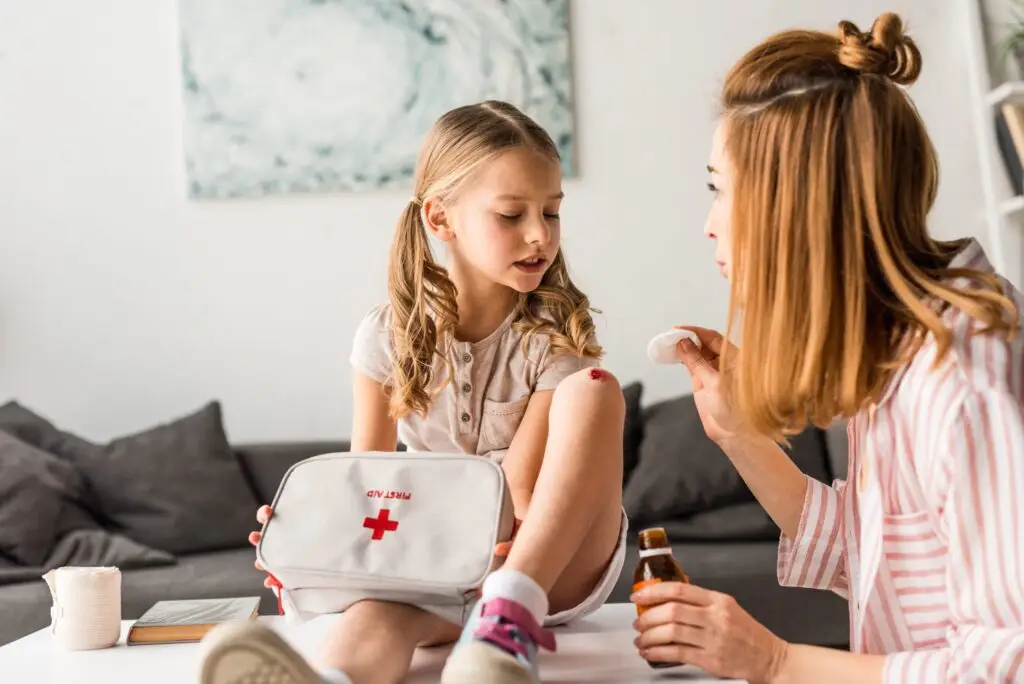 A woman demonstrates how to teach first aid in a fun way by gently cleaning a young girl's knee wound with a cotton pad, while holding a bottle of antiseptic. The girl, sitting with intrigue and a first aid kit on her lap in the cozy living room, smiles as she learns through care and attention.