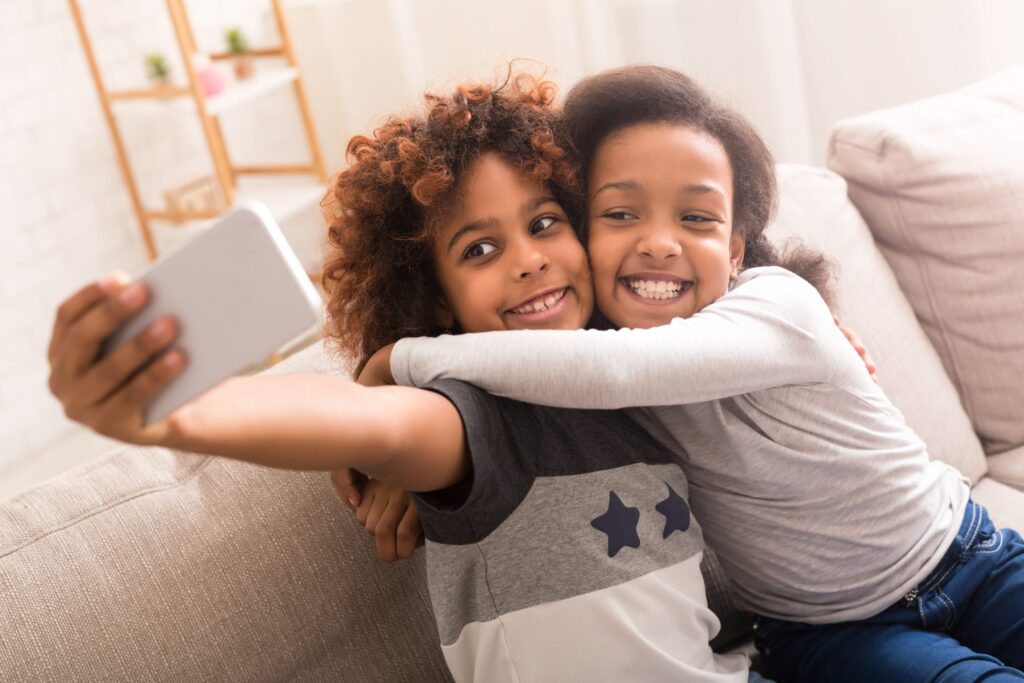 Two children sit on a couch, grinning as they take a selfie with a smartphone. One child, in a star-patterned shirt and curly hair, beams while the other, wearing a light top, hugs them. It's important to talk about social media safety for kids to keep moments like these secure and joyful.