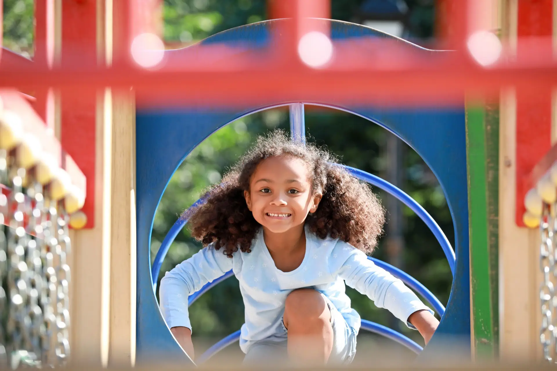 A young girl with curly hair and a blue shirt smiles as she crouches on the colorful, safe playground equipment. Bright sunlight highlights her joyful expression, and the background is blurred with greenery.