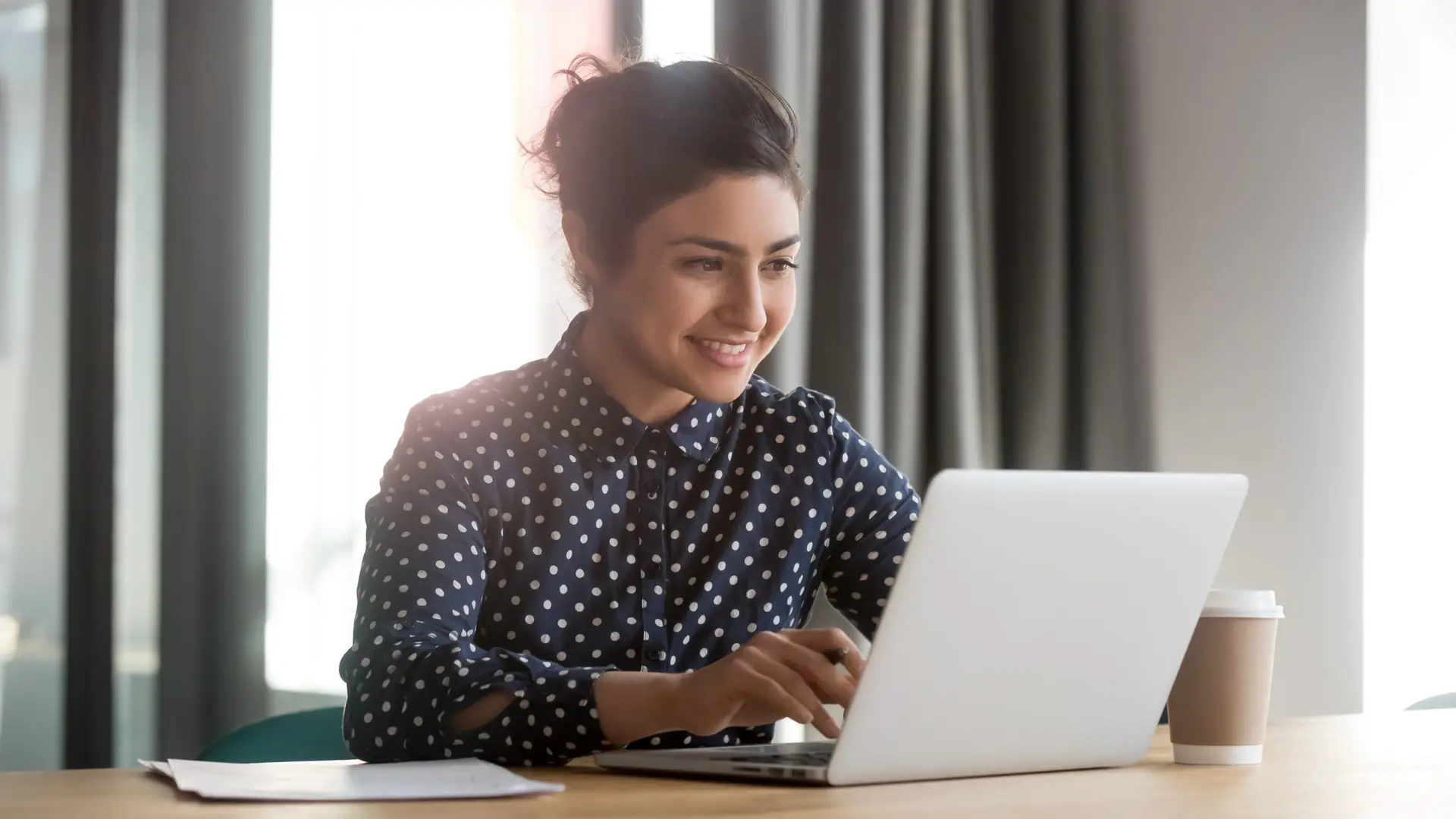 A woman in a polka dot blouse is smiling while using a laptop at a table, researching "How Do I Know a Website Is Credible?" A coffee cup and paper are next to her. Natural light filters through curtains in the background.