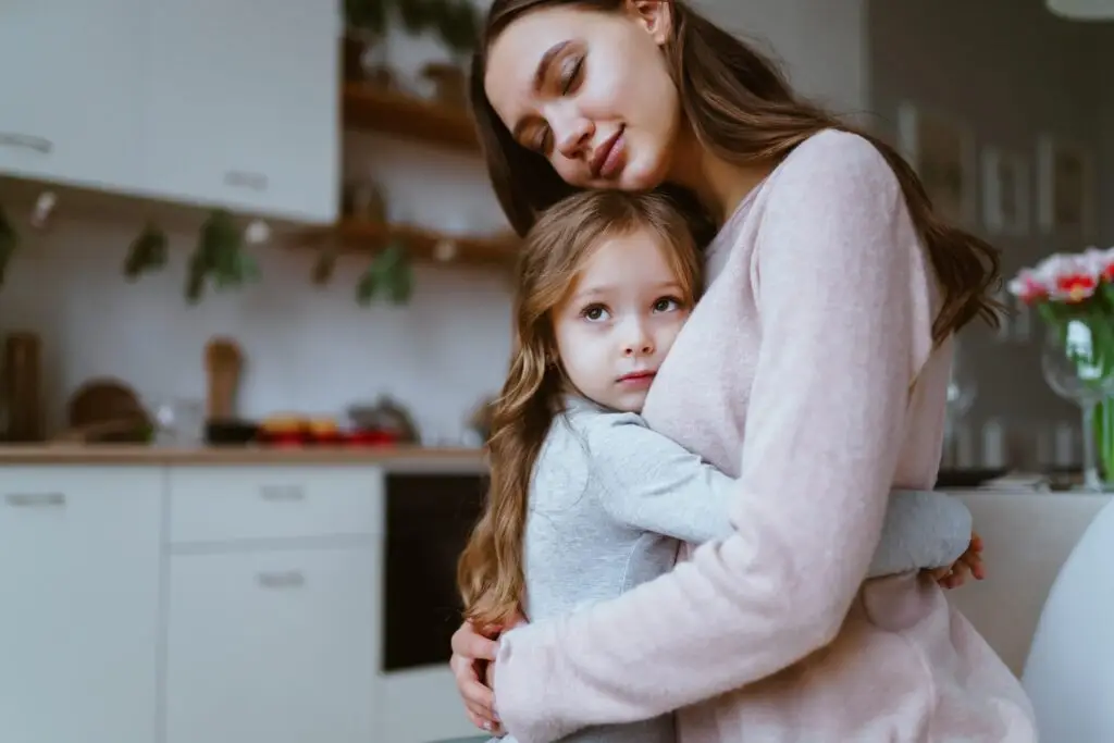 In a cozy kitchen, a woman and a young girl share a loving embrace, underscoring the warmth and patience needed to support each other. The woman gently rests her head on the serene girl's, as if to reassure her that even when one cries over everything, solace can be found in tender moments.