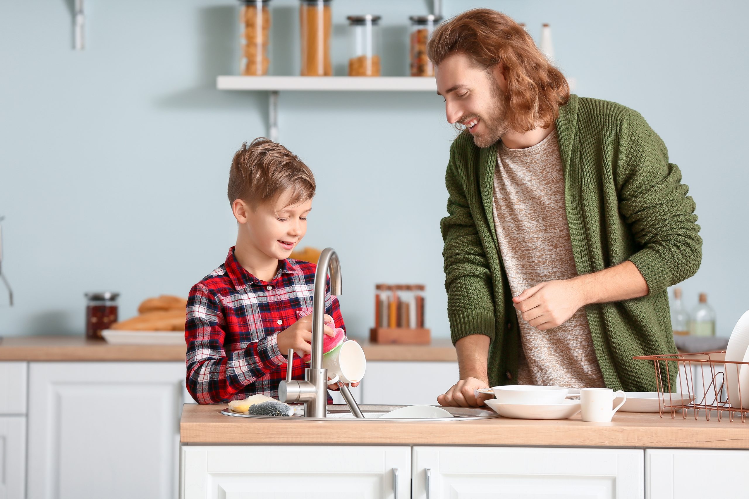 A man and a boy happily wash dishes together in a modern kitchen, following Troomi’s Handy List of Chores for Kids by Age. The man wears a green cardigan, and the boy sports a red plaid shirt as they stand at the sink with shelves and jars in the background.