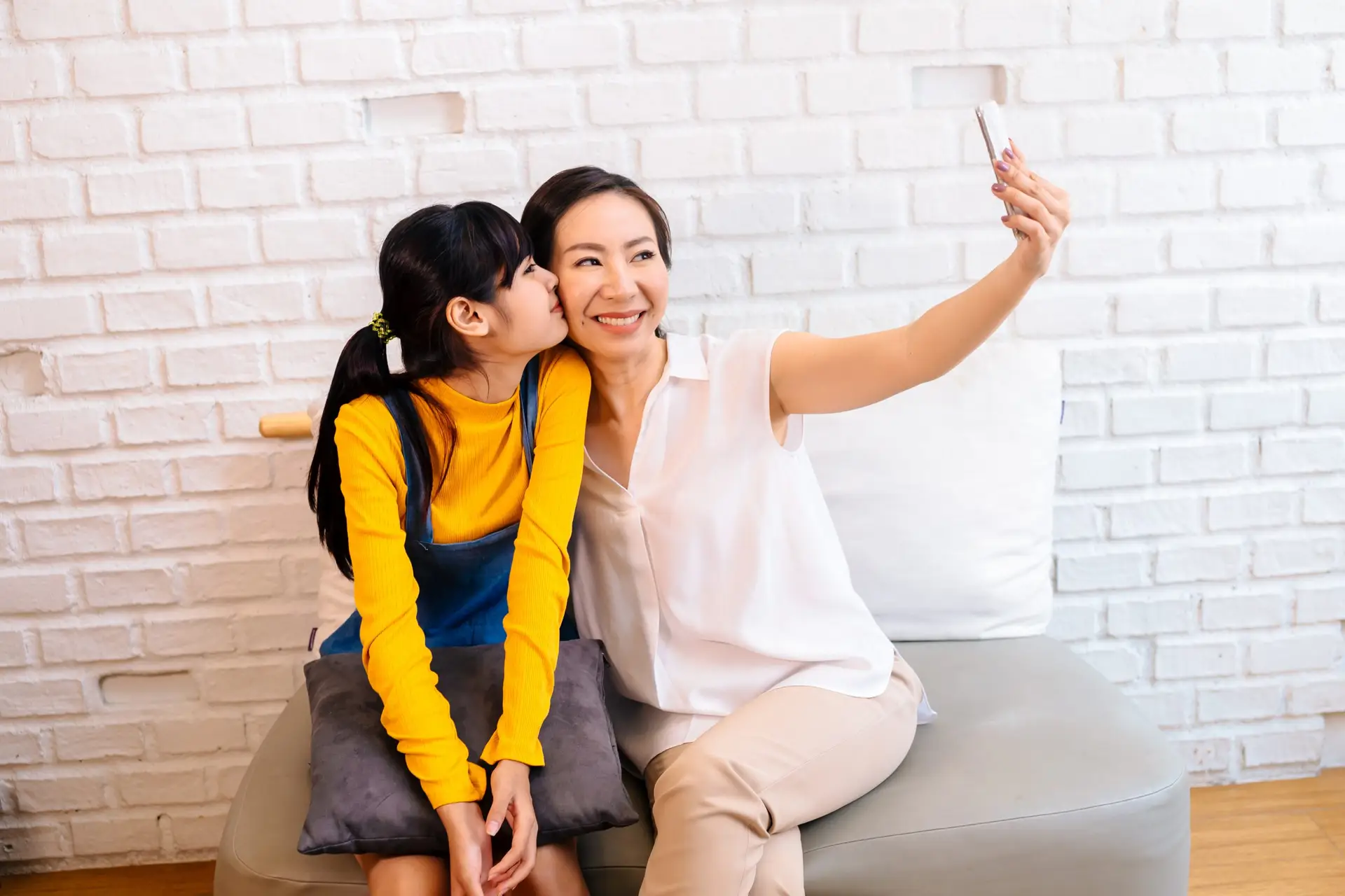 A woman in a white blouse takes a selfie with her smartphone, smiling, as she sits on the couch. A young girl in a yellow top and denim overalls kisses her cheek. The cozy scene reflects how online communities can bring joy to parents and families alike against a white brick wall backdrop.