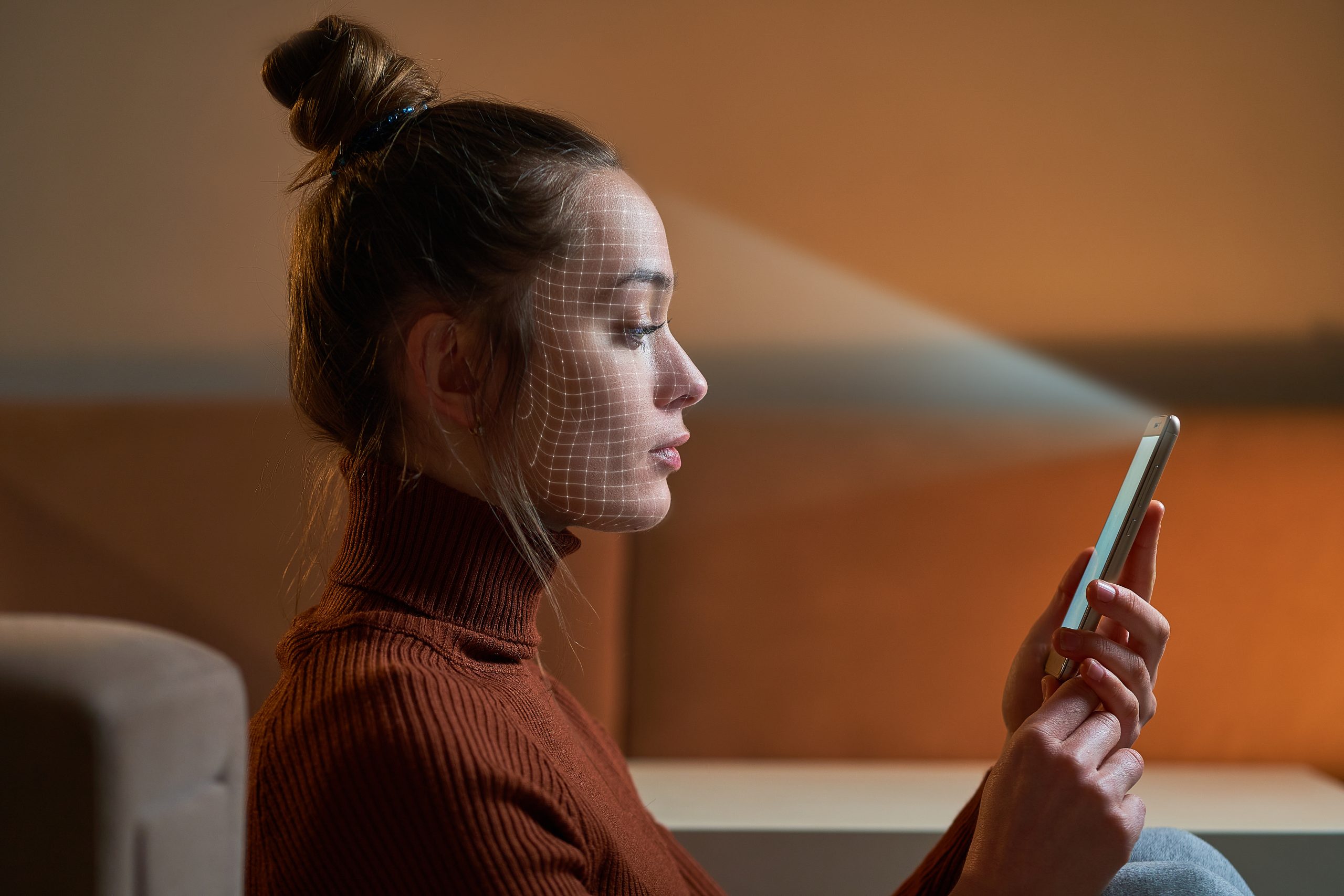 A woman in a brown turtleneck demonstrates how facial recognition works on her smartphone. A digital grid pattern is projected on her face, highlighting the scanning process. She sits indoors, against a neutral background, illustrating the seamless integration of technology into daily life.