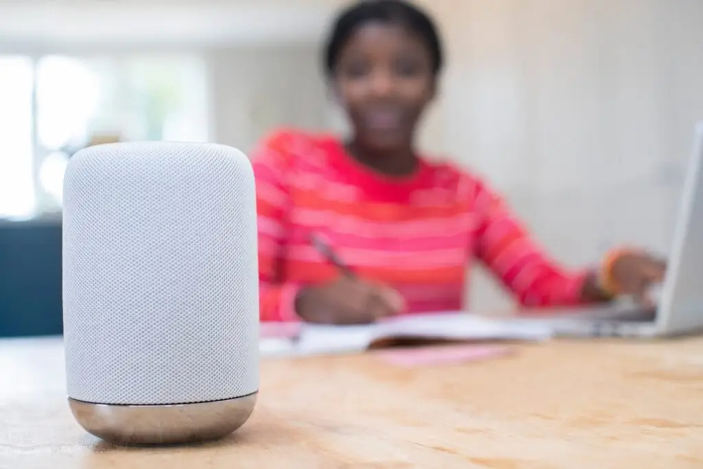 A smart speaker is placed on a wooden table in the foreground. In the background, a person wearing a red striped shirt uses a laptop and writes in a notebook, slightly out of focus. The setting appears to be a home or office, prompting the question: Are smart speakers really safe for children?