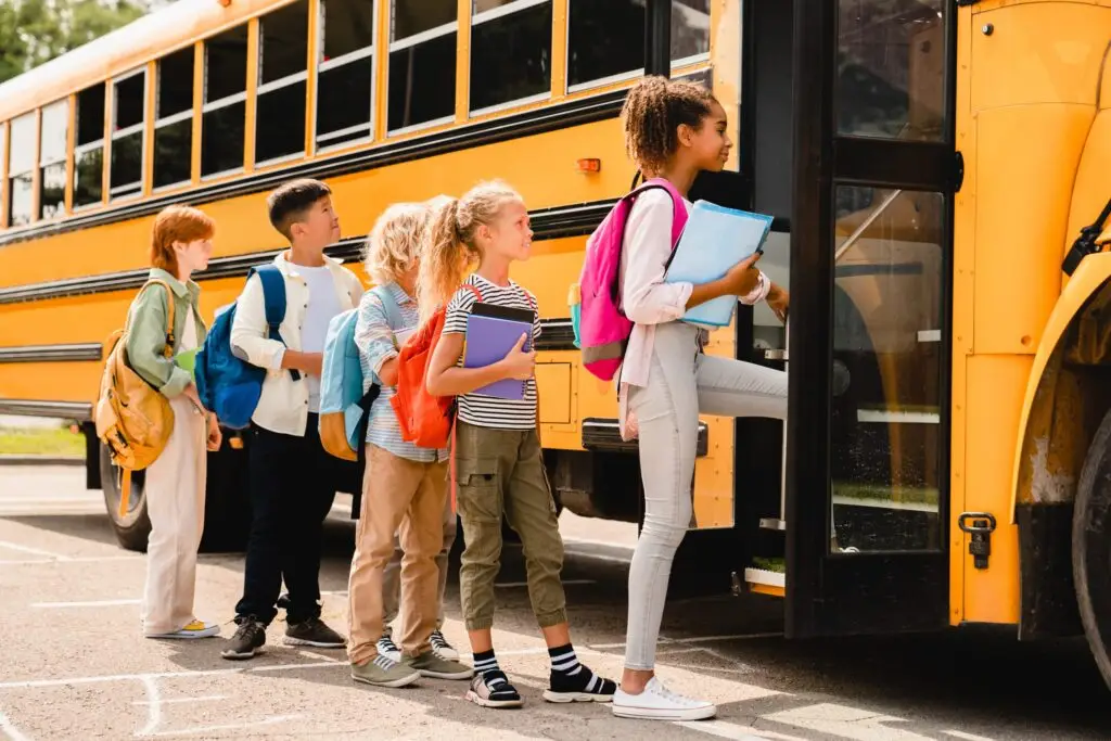 A group of children with backpacks stands in line for the yellow school bus, eager yet mindful. Clutching their notebooks and dressed casually, they illustrate School Bus Safety for Students: Three Trusty Tips, as they wait in the parking lot under a clear sky.