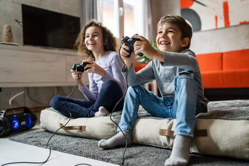 Two children, a girl and a boy, are sitting on a large cushion in a cozy living room. Smiling and focused, they're playing video games with controllers in hand. The modern decor features a TV and gaming console, reminding parents to consider five online gaming safety tips for your kids.