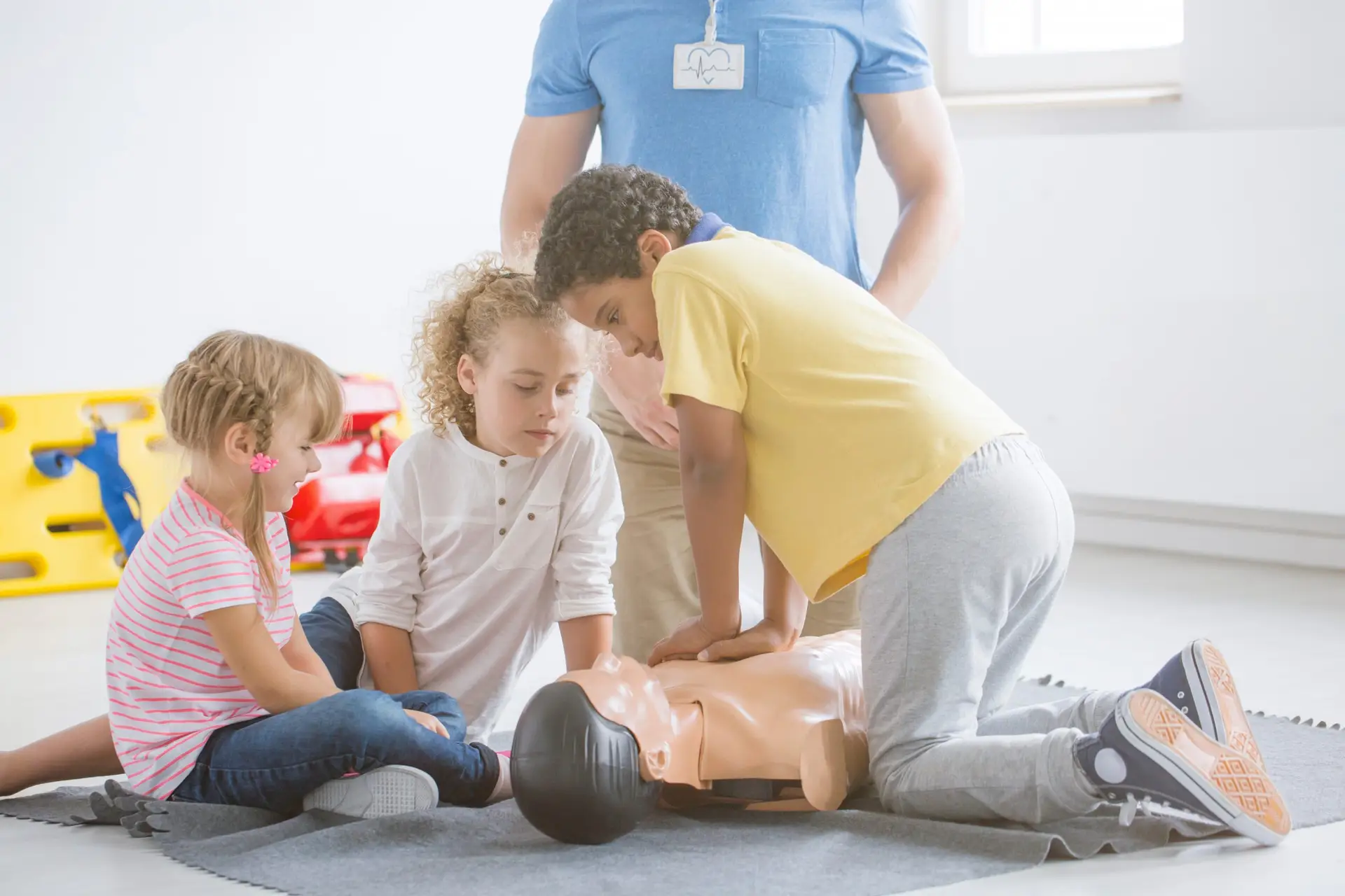 In a well-lit room with colorful furniture, three little lifesavers engage in CPR training for kids. The boy in the yellow shirt performs chest compressions on a manikin under adult supervision, while the other two children watch intently.