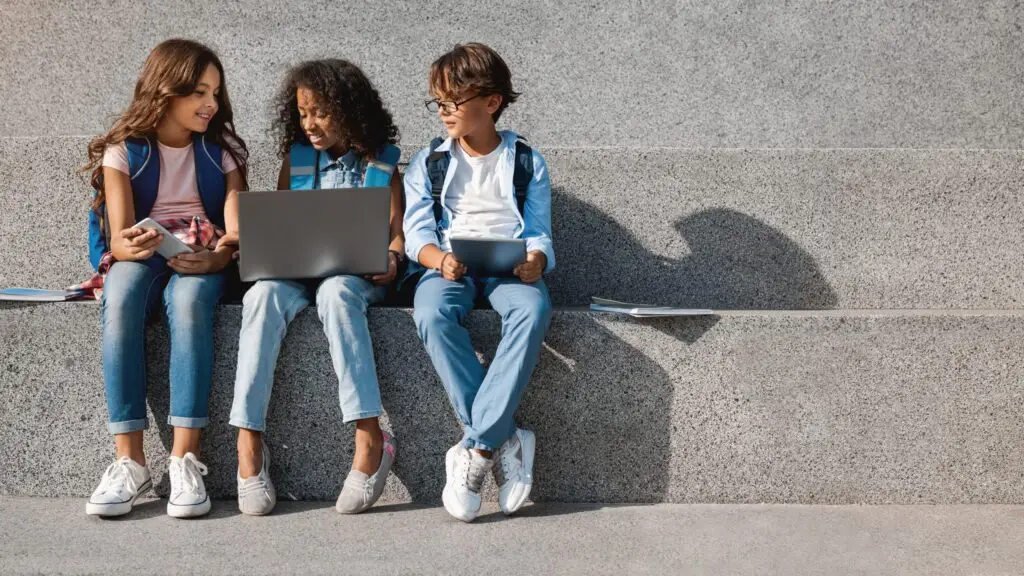 Three children sit on concrete steps with backpacks. One girl holds a phone, another uses a laptop, and a boy explores tablet apps designed for stalkerware removal. They're smiling and engaged in their activities, enjoying the sunny day as they learn what your family needs.