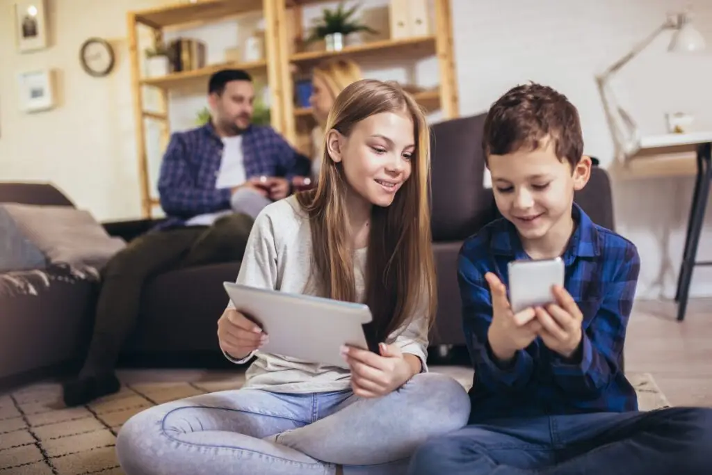 Two children sit on the floor, smiling and using a tablet and smartphone. In the background, two adults on a couch are engaged with a phone and a book, perhaps pondering, "Does technology make parenting harder?" The room is cozy with shelves and a lamp.