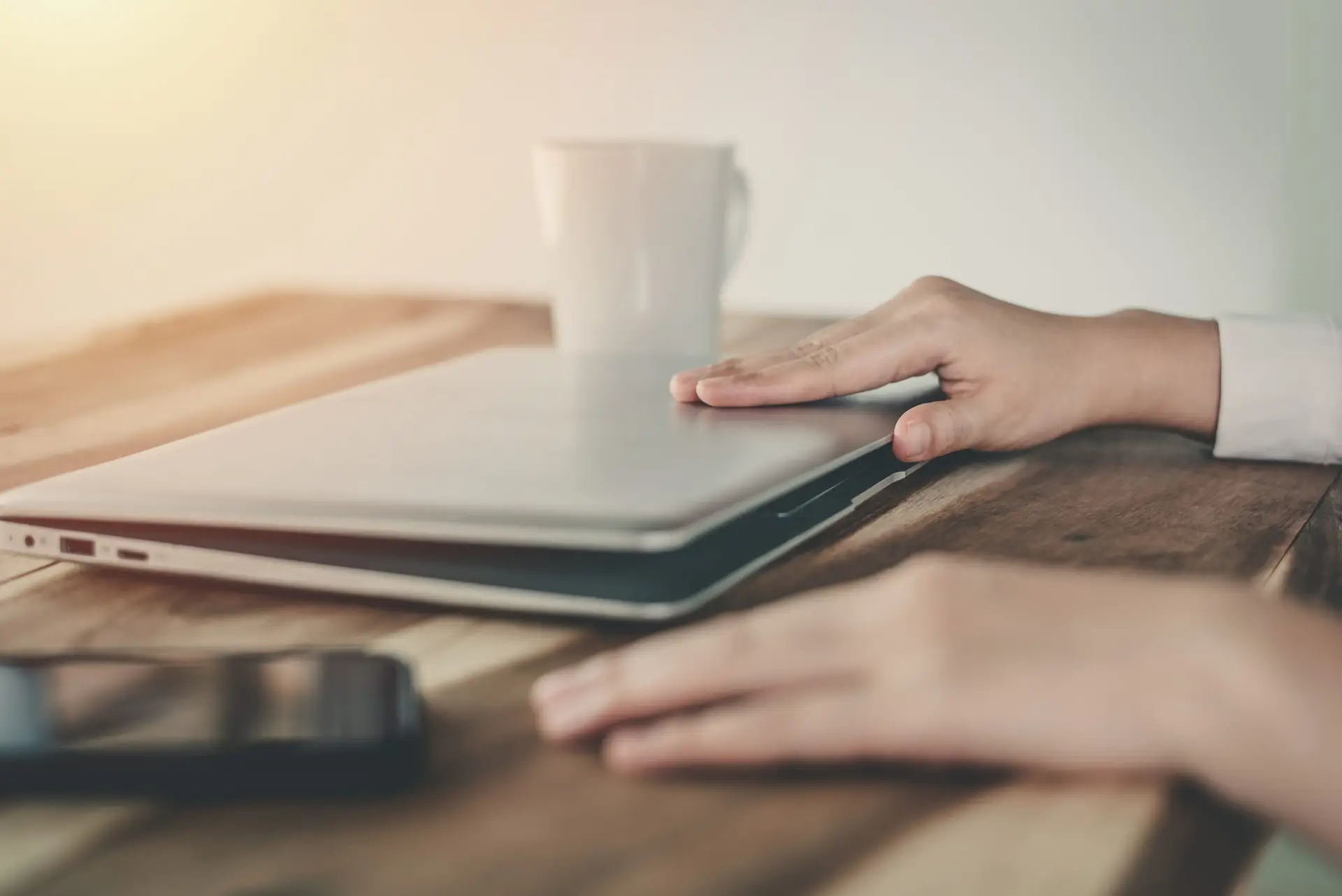 A person gently closes a silver laptop on a wooden desk, perhaps having just read an article titled "What Parents Need to Know About the Platform OnlyFans." Nearby, a smartphone and a white coffee mug are visible. Soft natural light illuminates the scene, creating a calm and serene atmosphere.