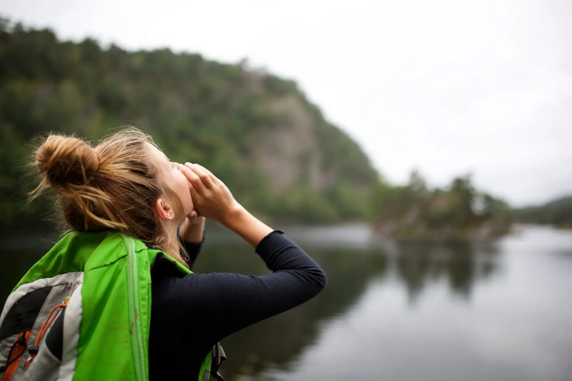 A person with a bun hairstyle and green backpack calls out towards a lake, surrounded by wilderness, as if echoing advice on what a missing child should do if they’re lost. The background features blurred trees and hills under a cloudy sky.