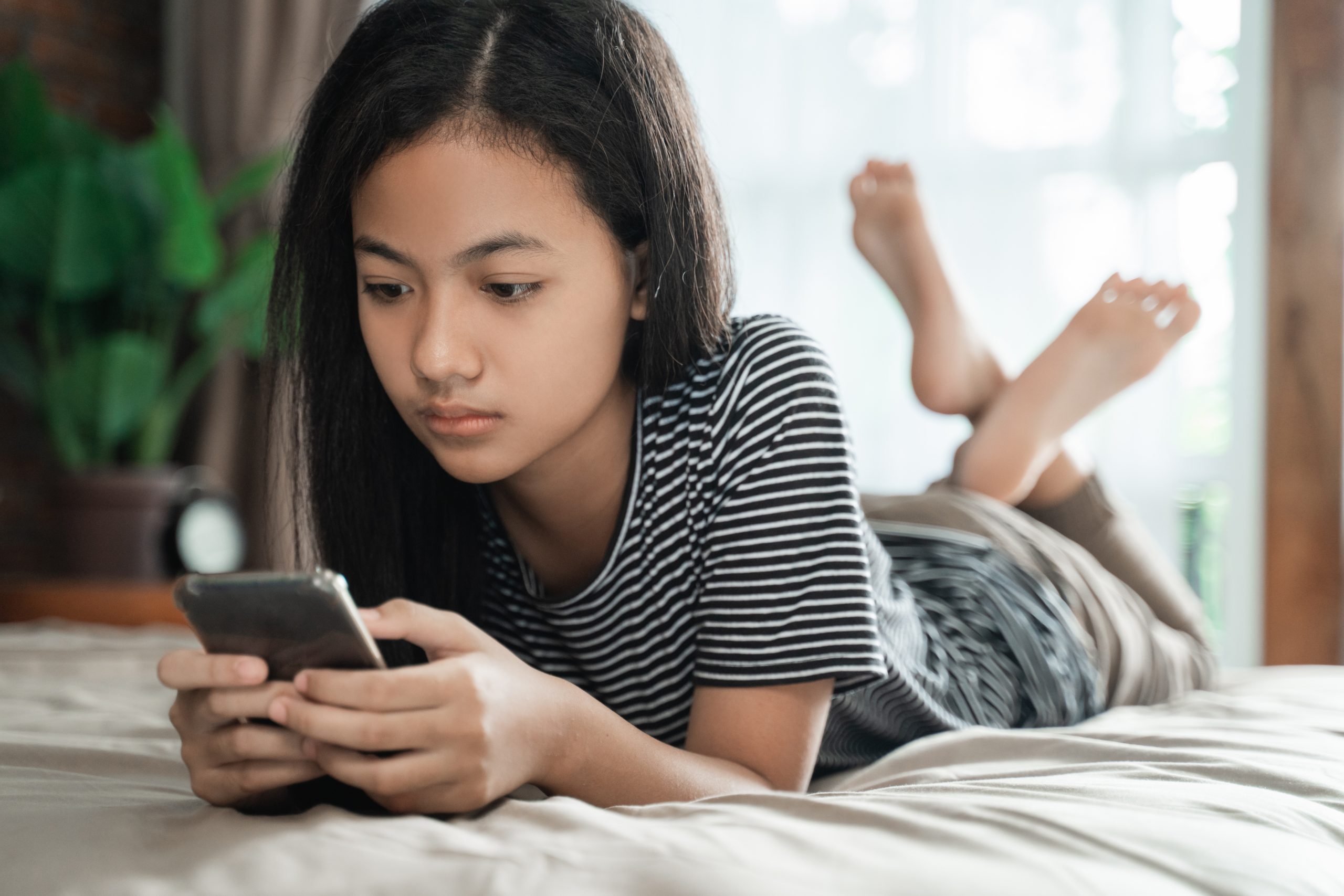 A girl in a striped shirt lies on her stomach on a bed, absorbed in her smartphone as she explores online friendships. Her legs are raised and crossed at the ankles, surrounded by soft natural light and green plants, creating a serene backdrop for this digital journey.