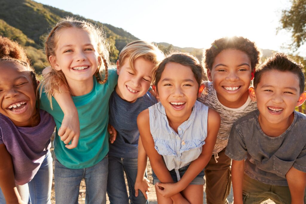 Six children stand closely together outdoors, smiling brightly at the camera. They appear happy and are dressed casually. The sun shines brightly in the background, highlighting the greenery and hills behind them, a perfect scene to ask: What is UNICEF doing to support such joyful childhoods?