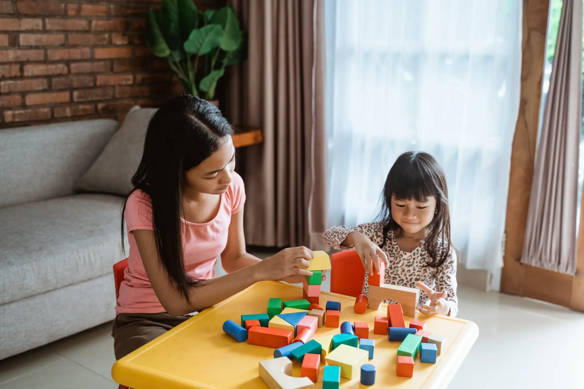 A woman and a child are seated at a yellow table, exploring colorful building blocks in their cozy living room—a perfect scene illustrating 6 reasons why your family should switch to a toy-sharing subscription. The woman sports a pink shirt, the child a patterned top, with a couch and large plant in the background.