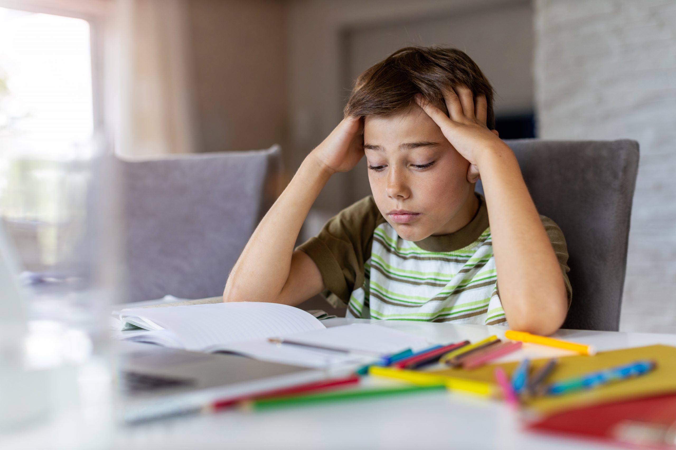 A young boy sits at a table, visibly frustrated, his head in his hands. Surrounded by open books and colored pencils, it's clear my child is struggling in school. What do I do to help him find focus and ease the stress of homework?