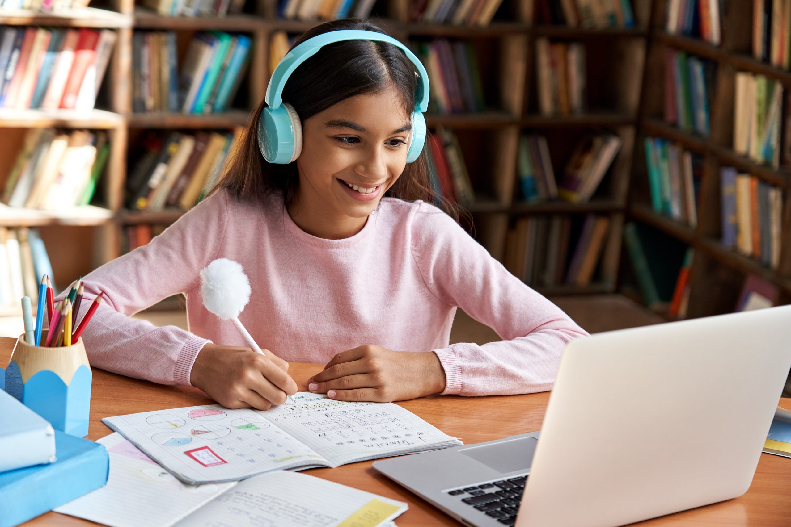 A young girl, aware of child protection laws, wears headphones as she sits at a desk in a library, smiling at her laptop while learning how to protect children online. She's holding a pen and writing in a notebook filled with colorful charts. Bookshelves are visible in the background.