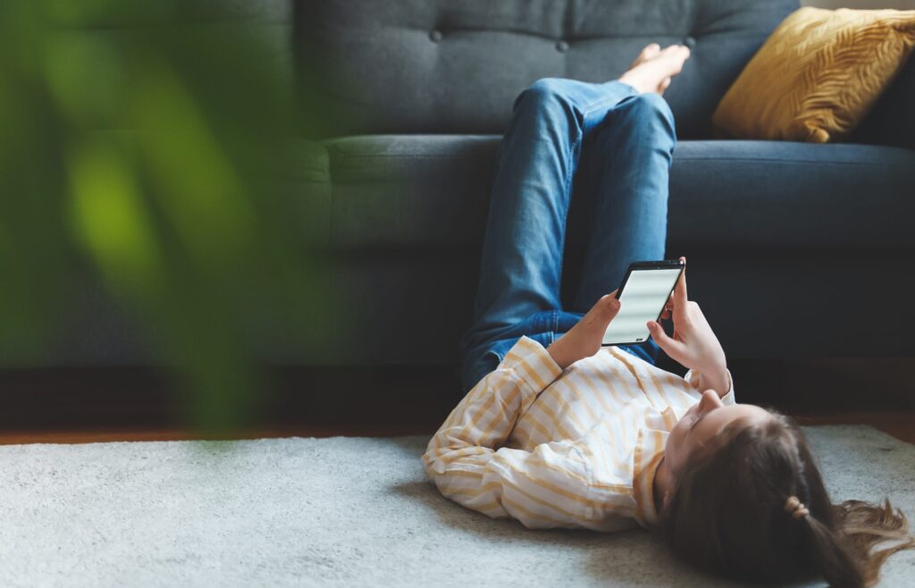 A person lies on the floor with their legs resting on a sofa, looking at a smartphone, pondering "Is Twitter Safe for Kids?" Dressed in a striped shirt and jeans, they relax amidst a green plant in the foreground and rest against a yellow cushion on the sofa.