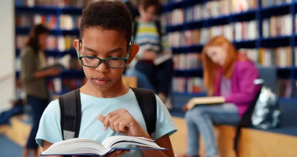 A child wearing glasses and a backpack reads a book in the library, perhaps diving into fanfiction stories. In the background, two others sit on steps reading, surrounded by shelves full of books. The atmosphere is quiet and studious—a perfect setting for exploration and what parents need to know.