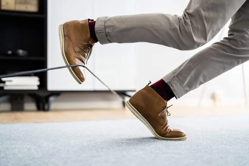 In an office setting, a person trips over a black cable, highlighting the importance of covering cables to prevent trip hazards. The image shows one leg raised, wearing light brown shoes and gray pants, as the precarious situation unfolds.