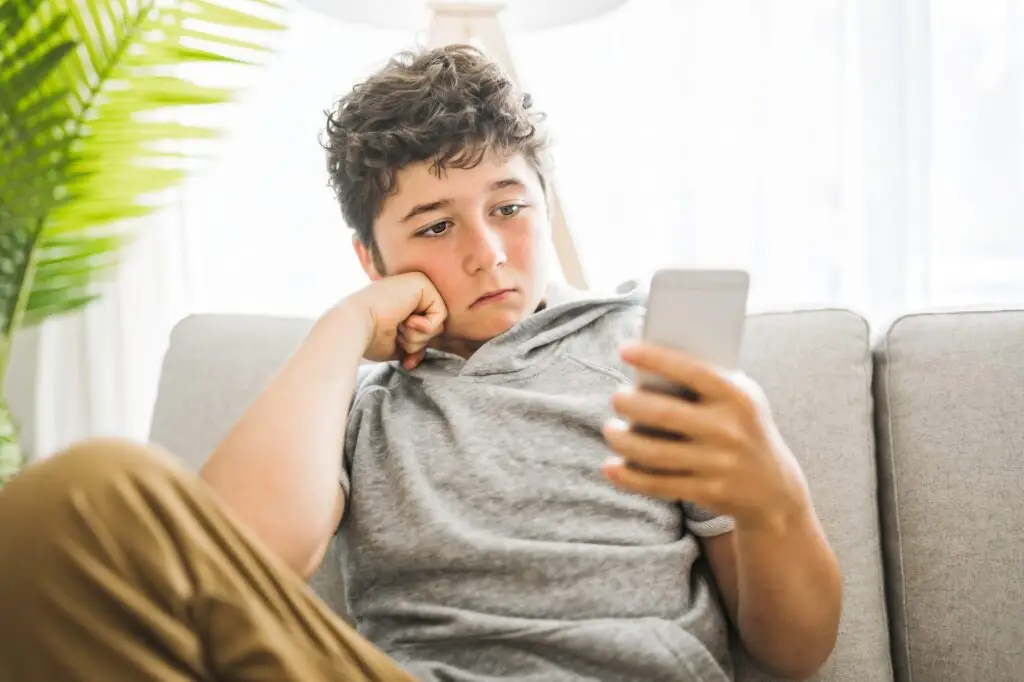 A boy with curly hair and a gray hoodie sits on a couch, gazing intently at a smartphone. He rests his head on his other hand, appearing thoughtful as if pondering questions like "Is Pinterest safe for kids?" A leafy plant adds a touch of greenery in the background.