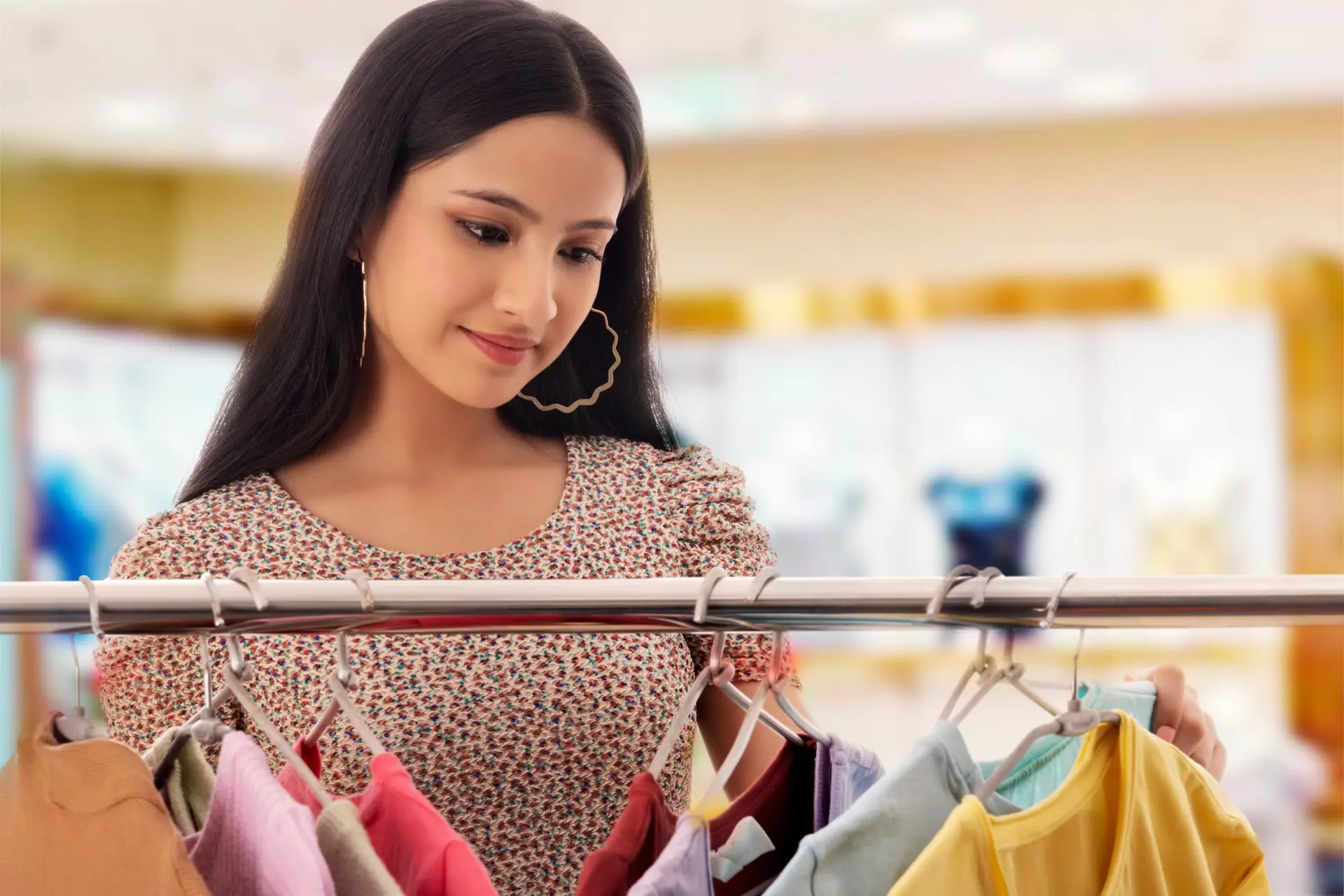 A woman with long dark hair, wearing a patterned dress and large hoop earrings, is browsing colorful clothes on a rack in a store. With the soft blur of the bright and inviting shopping atmosphere, it's easy to imagine teens exploring their style as they discover ten ways to make money.