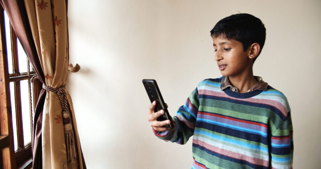 A young boy holding a smartphone looks at the screen with interest, perhaps wondering, "Is Reddit Safe for Kids?" He is wearing a colorful striped sweater and stands near a window with beige curtains in a room with light-colored walls. Parents might want to know more about his online activities.