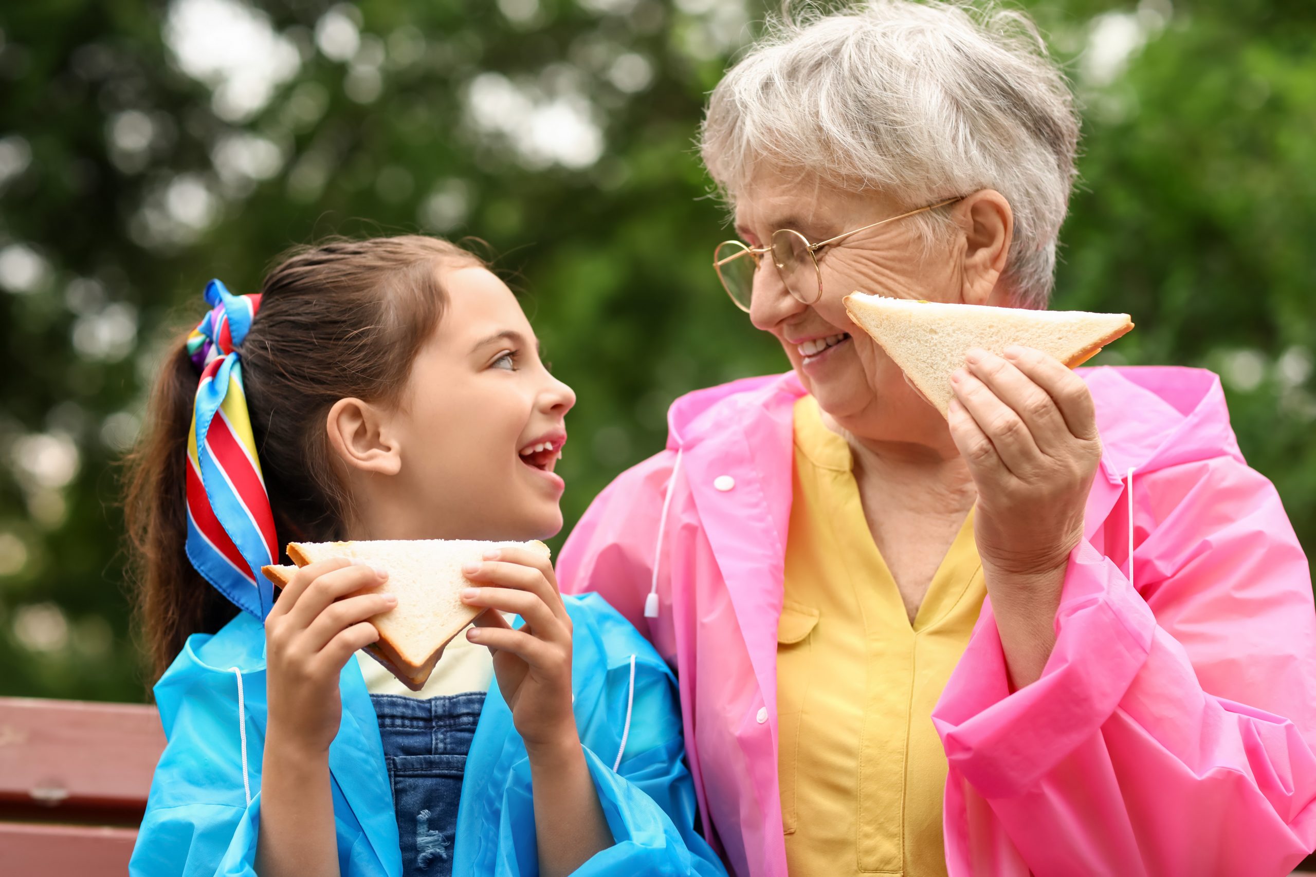 A grandmother and granddaughter sit on a bench, smiling at each other while holding sandwiches. The grandmother, in a pink raincoat and glasses, carefully considers how to help friends with food allergies as her granddaughter, dressed in blue with colorful ribbons, eagerly takes a bite.