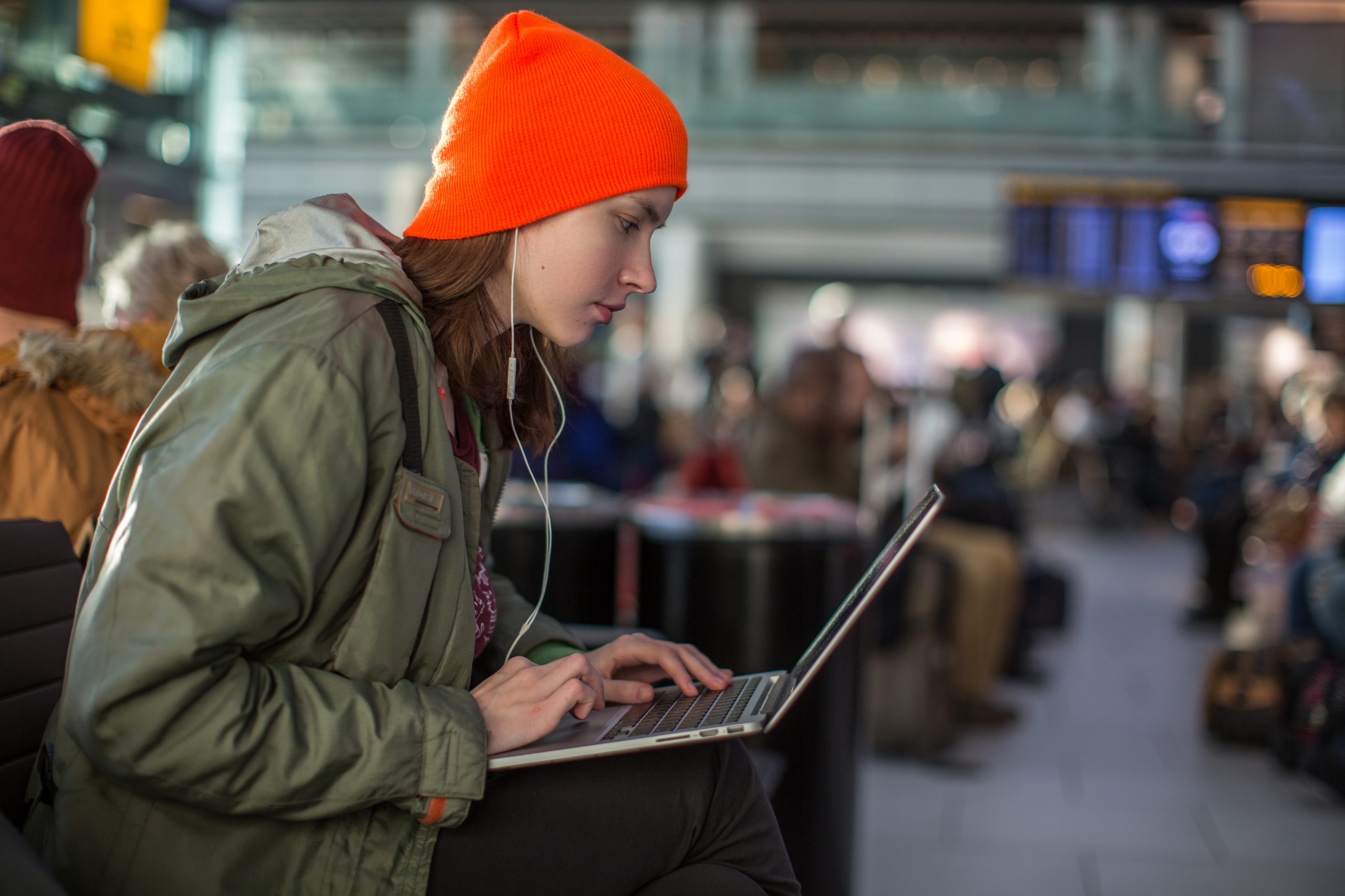 A person in an orange beanie and green jacket is intently focused on learning how to stay safe on public Wi-Fi while using a laptop in a bustling airport terminal, surrounded by other travelers and blurred departure screens in the background.