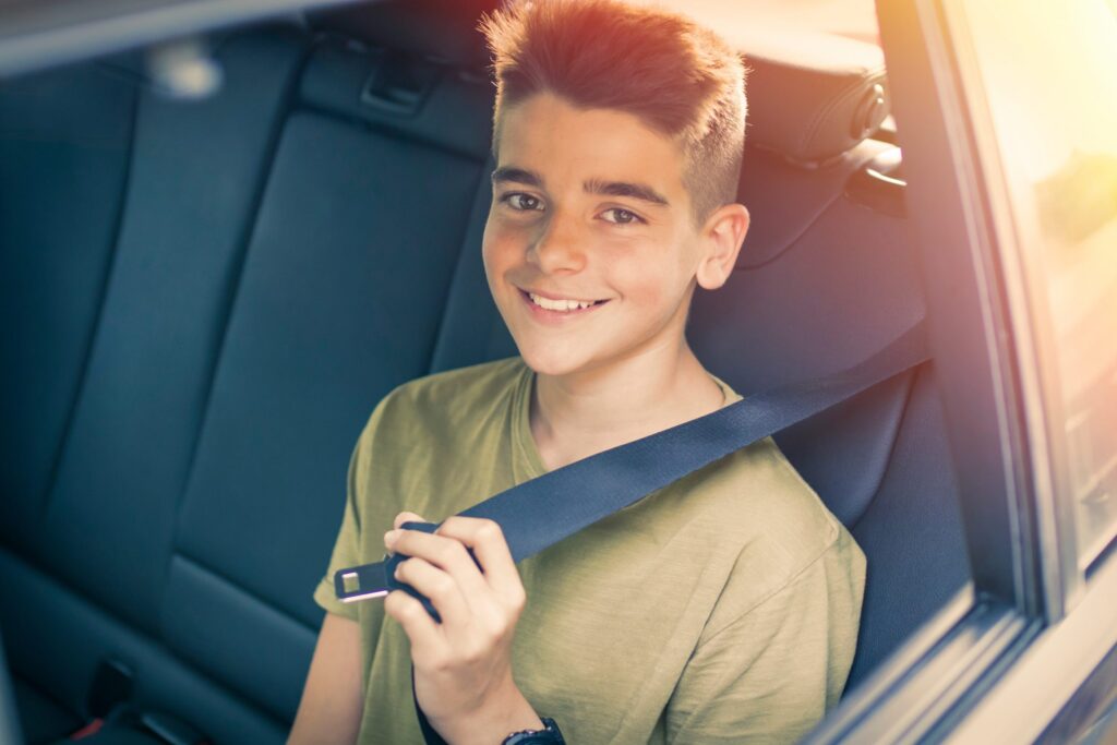 A young person with short hair is sitting in the backseat of a car, smiling as they prioritize safety by fastening their seatbelt. Sunlight filters through the window, casting a warm glow inside the car.