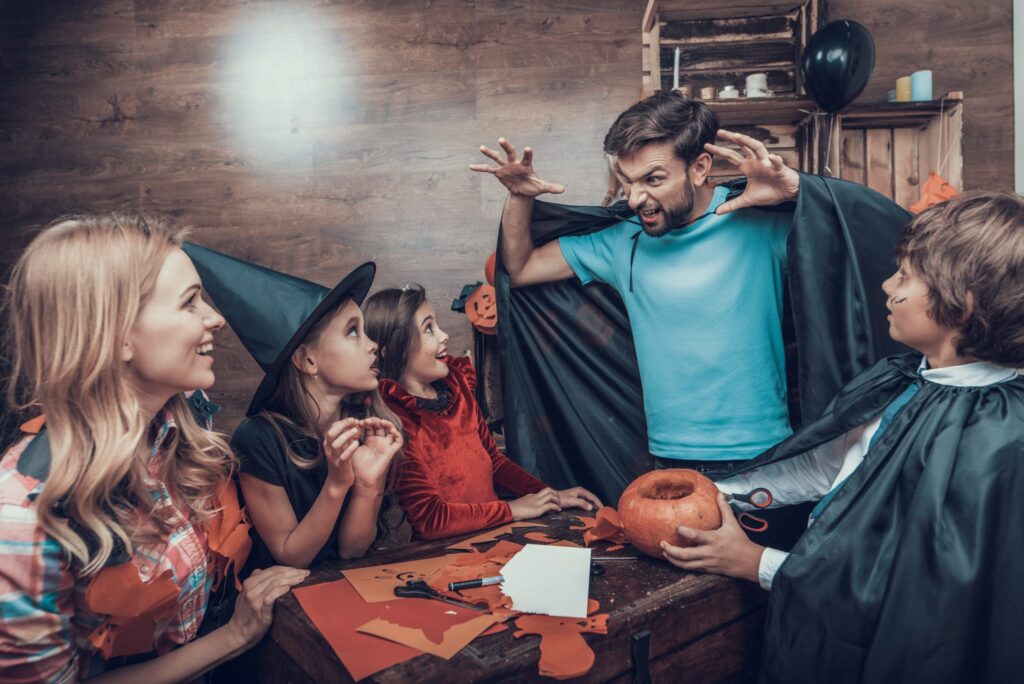 A man in a blue shirt and black cape playfully poses as a monster, surrounded by children in DIY Halloween costumes. They gather around a table with pumpkin crafts and decorations. The scene is cheerful and festive.