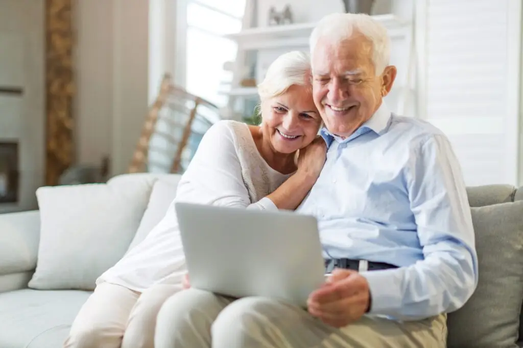 An elderly couple sits on a couch, smiling as they explore technology together on a laptop. The woman leans affectionately on the man's shoulder. The room is well-lit and cozy, with soft furnishings and a large window in the background.