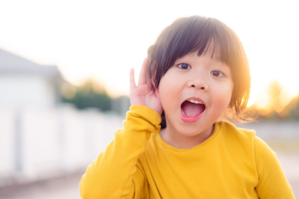 A young child in a yellow shirt holds one hand to their ear and listens intently, embodying the essence of listening for kids. They stand in an outdoor setting with a bright, blurred background.