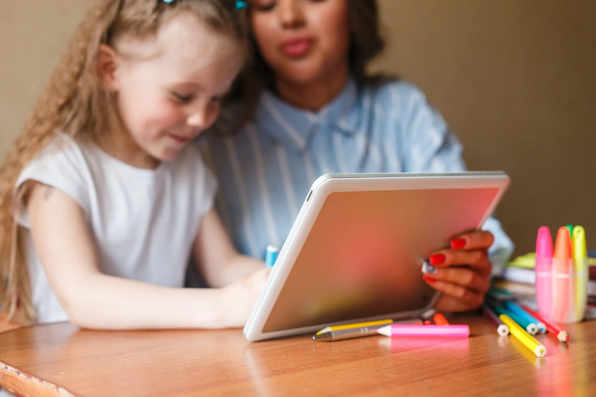 A young girl and a woman sit at a table filled with colorful markers and pencils, engaging in a time management activity on a tablet. The girl is focused on the screen as the woman looks on, smiling.