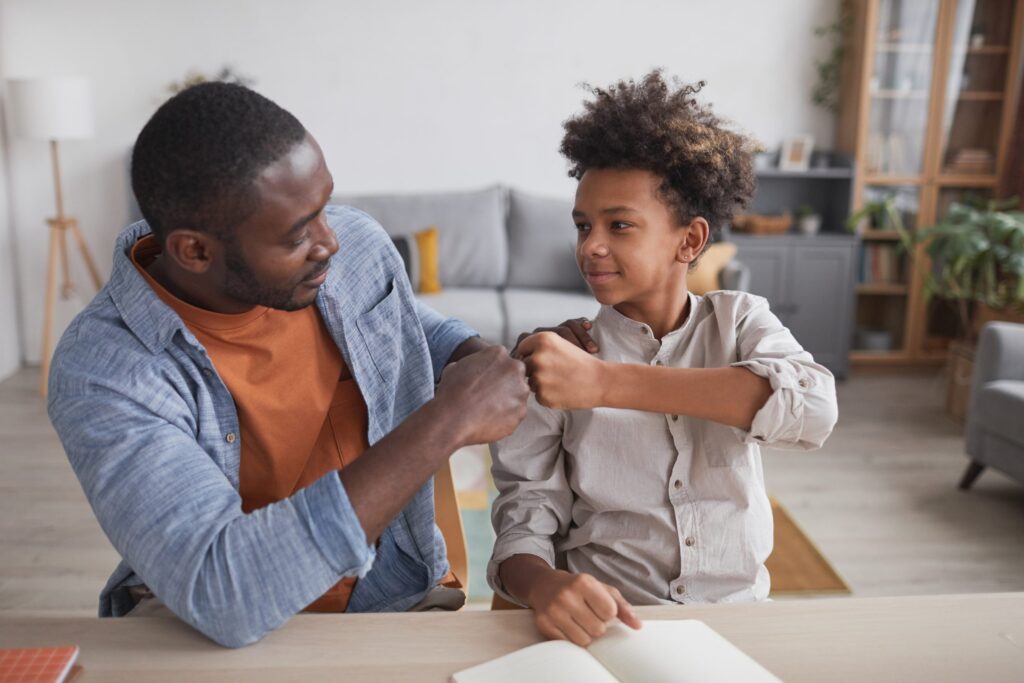 A man in a blue shirt shares a fist bump with a curly-haired child at the table, both smiling warmly in their cozy living room. It's moments like these that underscore the importance of understanding how to protect your child from sexual abuse, ensuring peace and security in their everyday life.