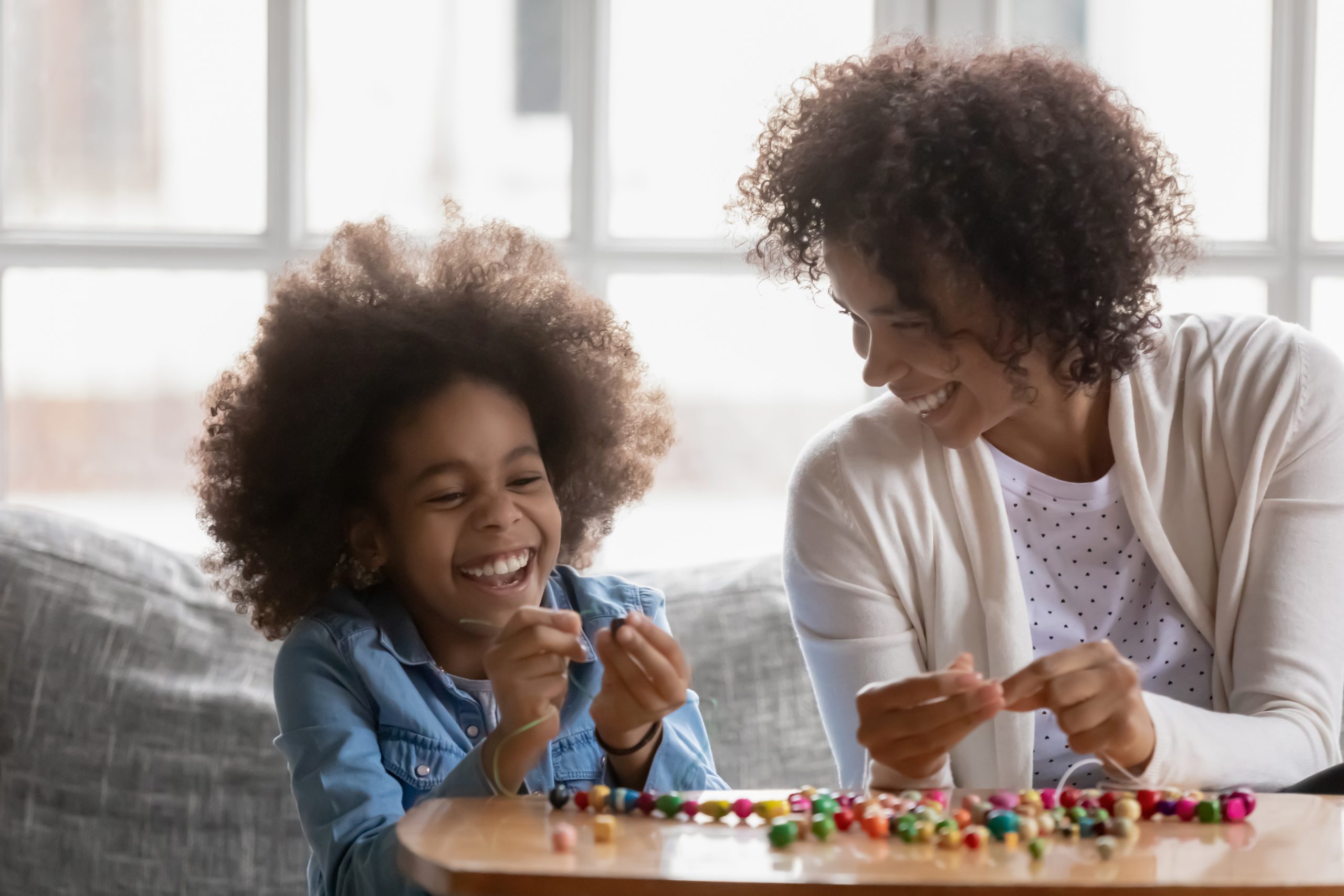 A mother and daughter sit at a table, smiling and laughing while threading colorful beads, showcasing a warm and joyful scene. This shared activity against a bright window backdrop highlights how technology can make parenting easier by fostering such cherished moments together.