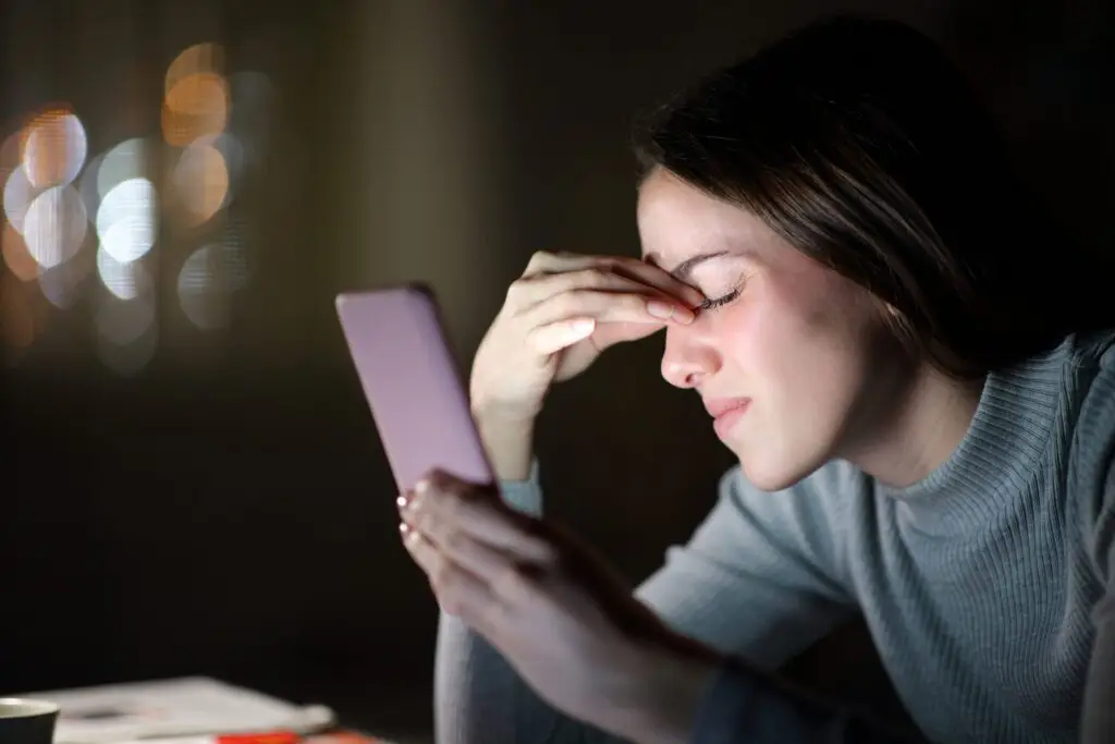 In a dimly lit room, a woman appears tired or stressed, holding a smartphone as the soft glow of its screen reflects on her face. Wearing a gray sweater, she rubs her forehead, seemingly burdened by concerns about cell phone radiation.