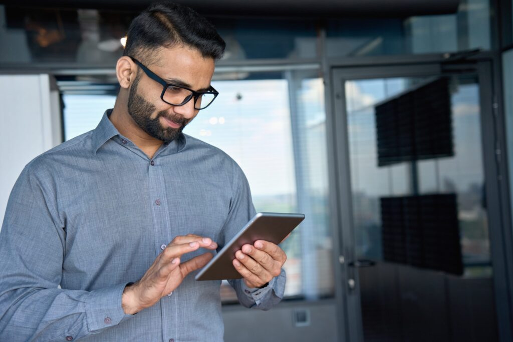 A man with glasses and a beard explores the latest tech gadgets for men on his tablet in an office with glass walls, bathed in natural light. He's dressed sharply in a gray shirt, fully engaged with the digital world at his fingertips.