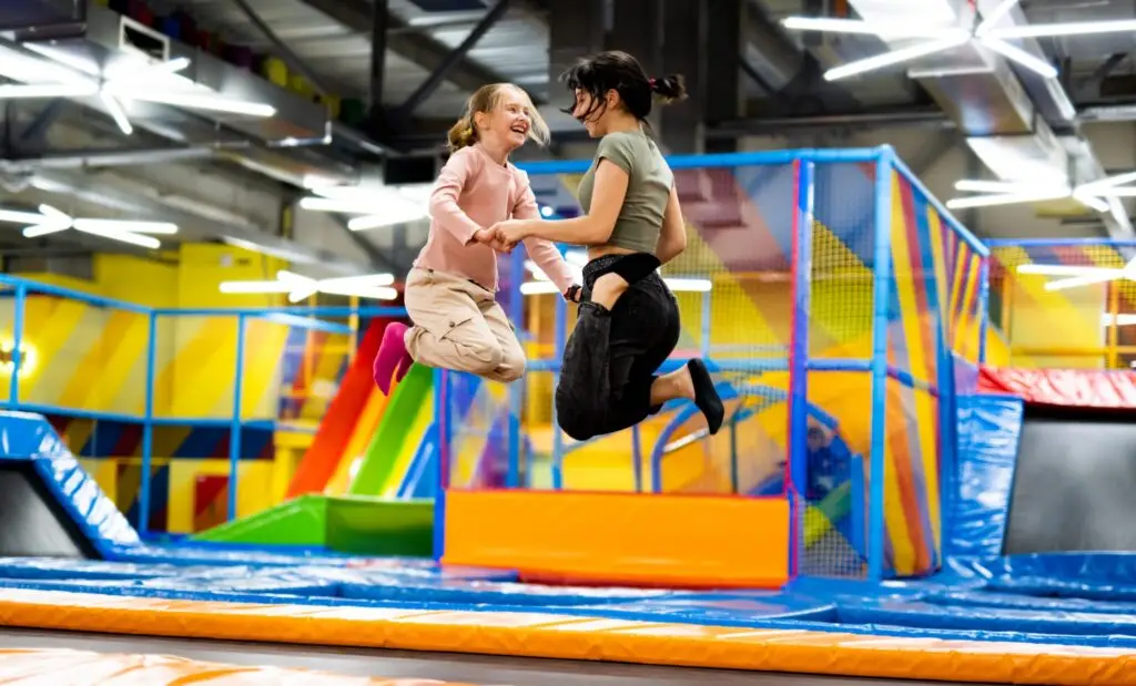 Two people are joyfully jumping on a trampoline in an indoor play center, enhancing their gross motor skills development. The colorful background features slides and climbing structures. They are holding hands and smiling, creating a lively and fun atmosphere.