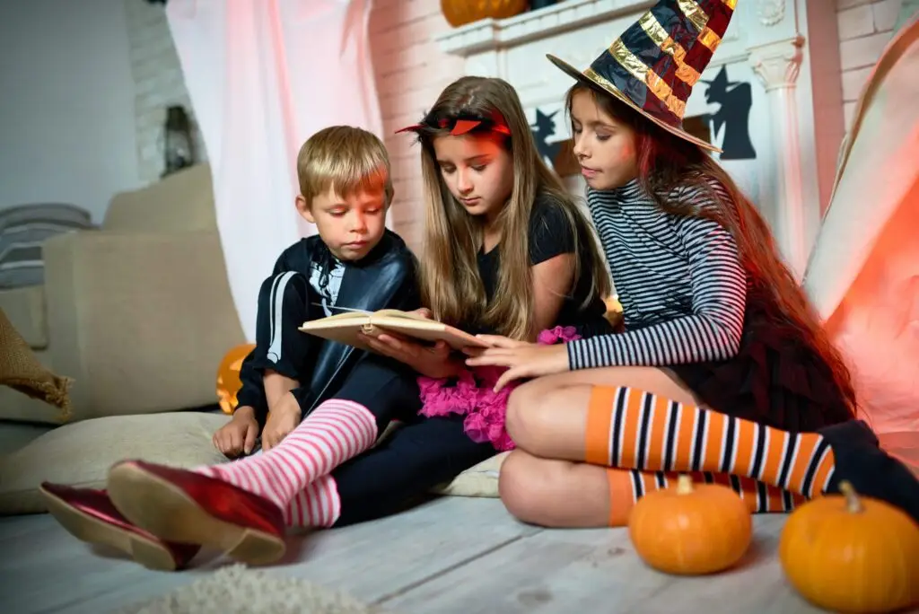 Three children in Halloween costumes sit on the floor reading "Scary Stories to Read Online." One wears a witch hat and orange-striped socks, another a devil headband, and the third a vampire cape. Small pumpkins are nearby, adding to their delight as they whisper about what Creepypasta might entail.