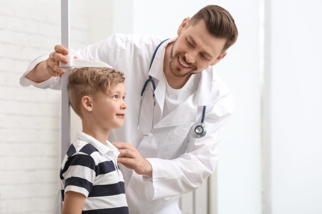 A doctor measures a young boy's height with a stadiometer. The smiling boy, in a striped shirt, stands still as the doctor, clad in a white coat and stethoscope, adjusts the tool. In the bright room, one might wonder: are tall children treated differently?