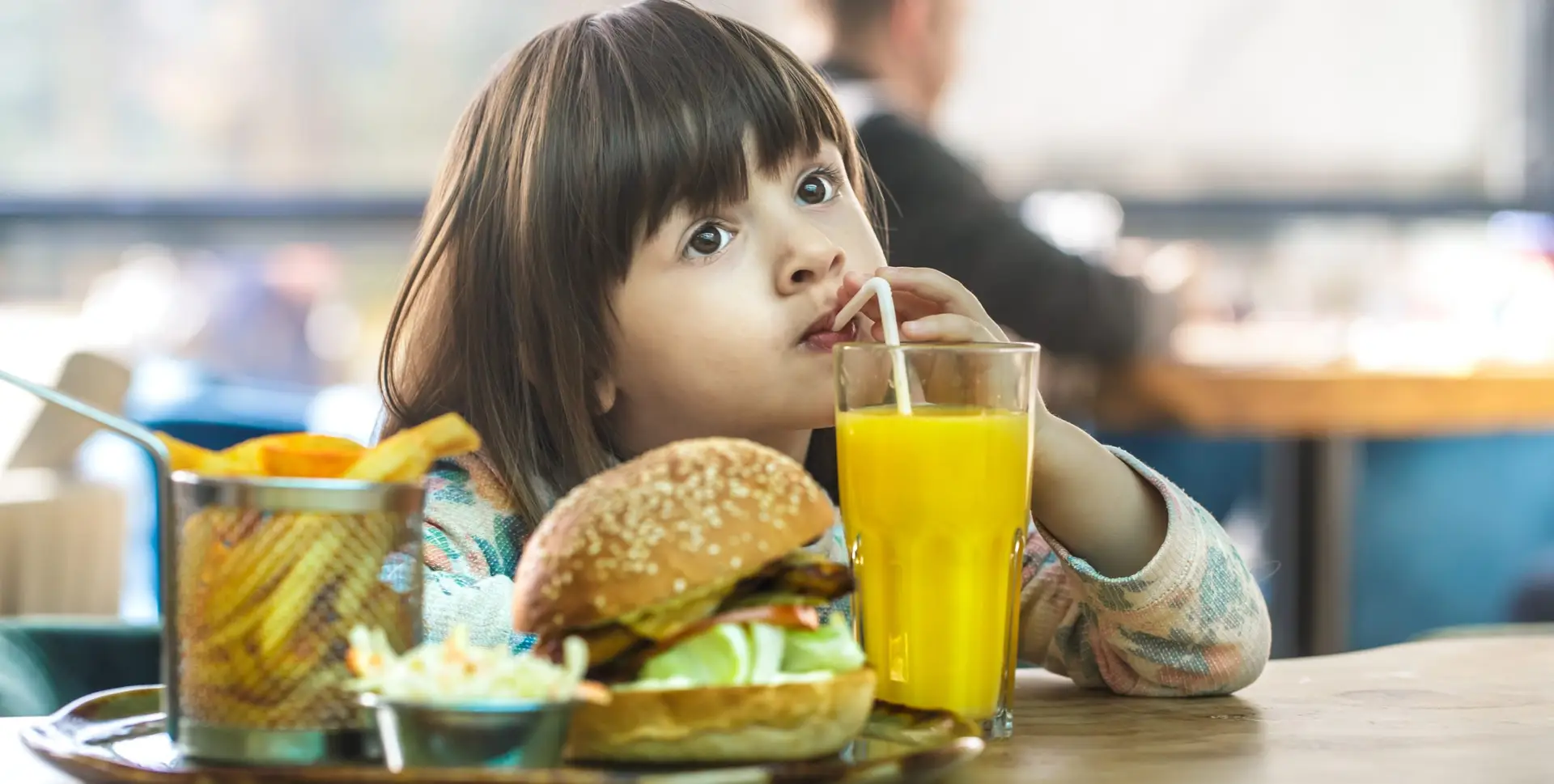 A young girl sits at a table in a restaurant, sipping orange juice through a straw. In front of her is a burger, a basket of fries, and a small dish of coleslaw. She looks thoughtful, perhaps pondering the allure of fast food that often captivates childhood cravings.