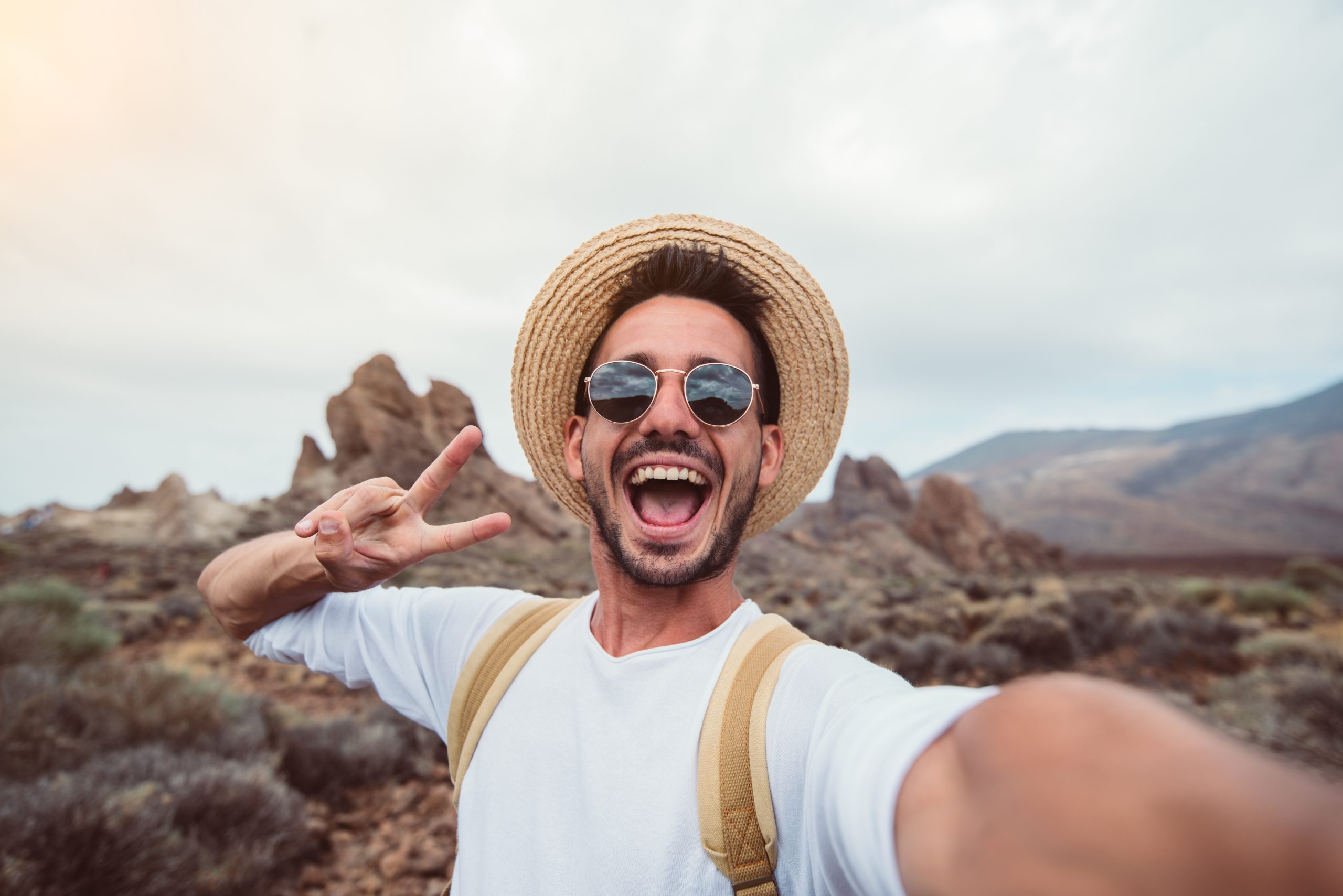 A person wearing sunglasses and a straw hat is taking a selfie in a rocky desert landscape, perhaps influenced by social media trends. They are smiling widely and making a peace sign with one hand against the cloudy sky, with mountains visible in the background.