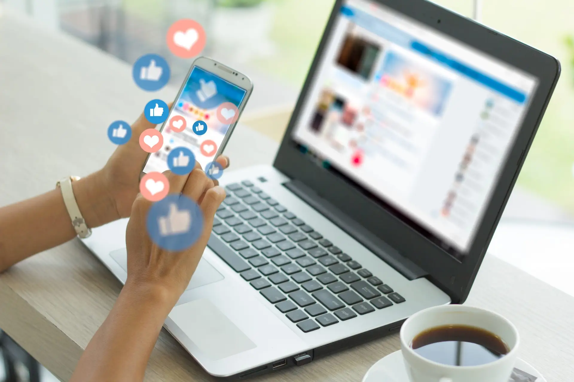 A person interacts with their smartphone, navigating a social media app displaying floating like and heart icons. Nearby, a laptop with the effects of social media on children open sits next to a cup of coffee on a light-colored table.