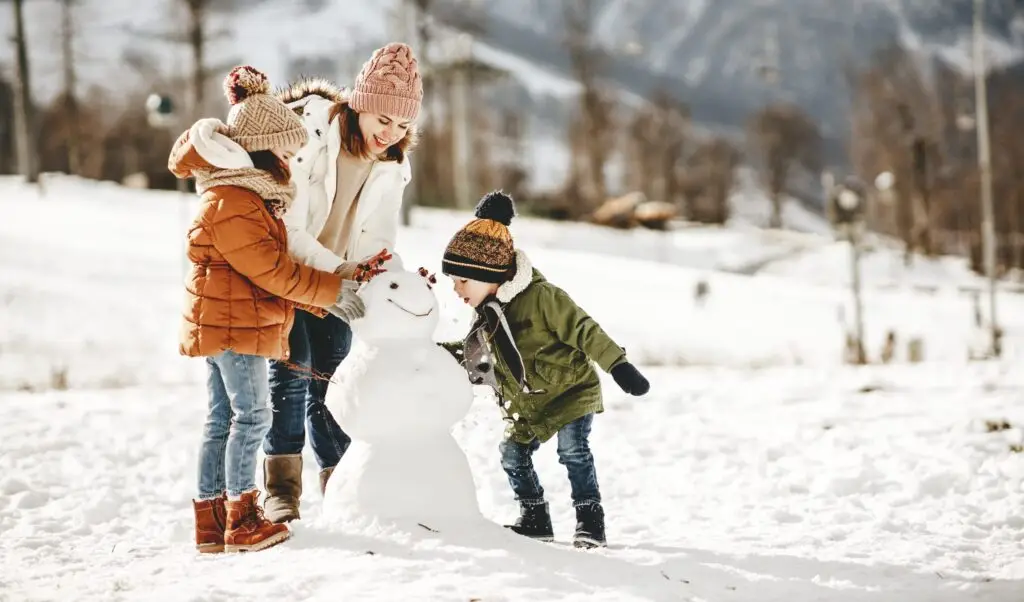 A woman and two children, bundled in warm clothing, are building a snowman in a snowy field. With snow-covered trees and mountains as their backdrop, the children smile as they add details to the snowman's face, exemplifying one of many winter joys while keeping Ten Safety Tips for the Winter Season in mind.