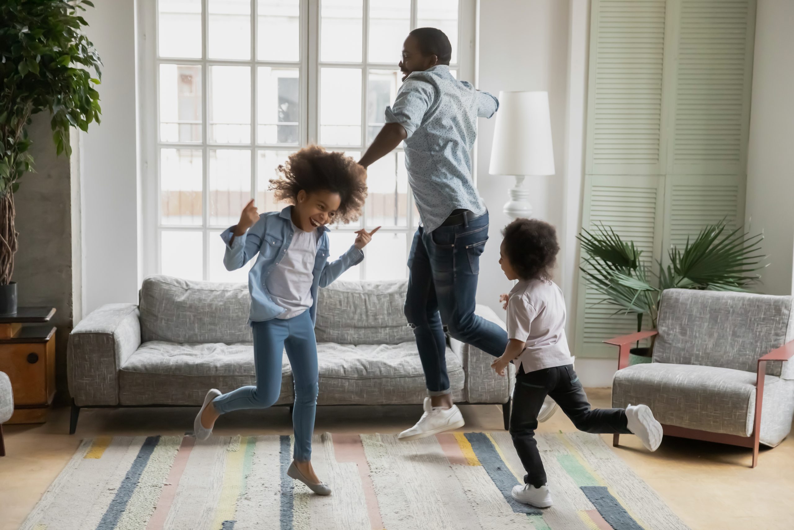 A joyful family dances together in a bright living room with large windows. Two kids and an adult, showcasing fine and gross motor skills, are mid-jump on a colorful striped rug, surrounded by cozy furniture and a potted plant. Everyone looks happy and energetic.