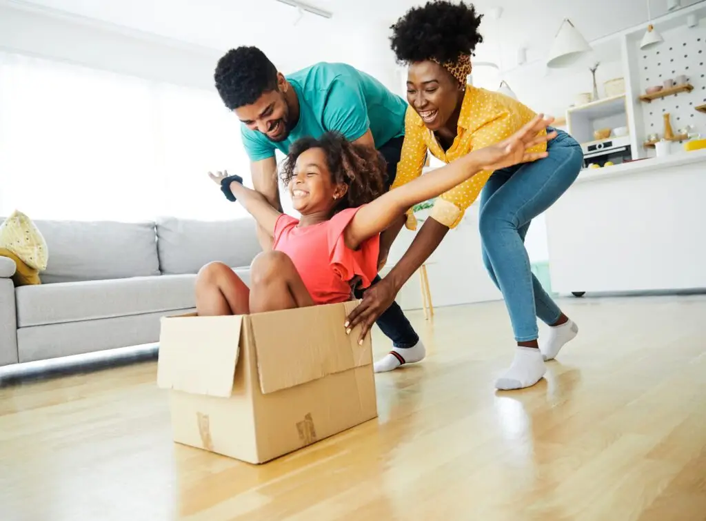 A joyful family in a bright living room enjoys some Family Fun. A child sits in a cardboard box with arms outstretched, pushed by two smiling adults, making weekend activities for families unforgettable. The scene conveys happiness and playful energy against a backdrop of a cozy sofa and kitchen area.