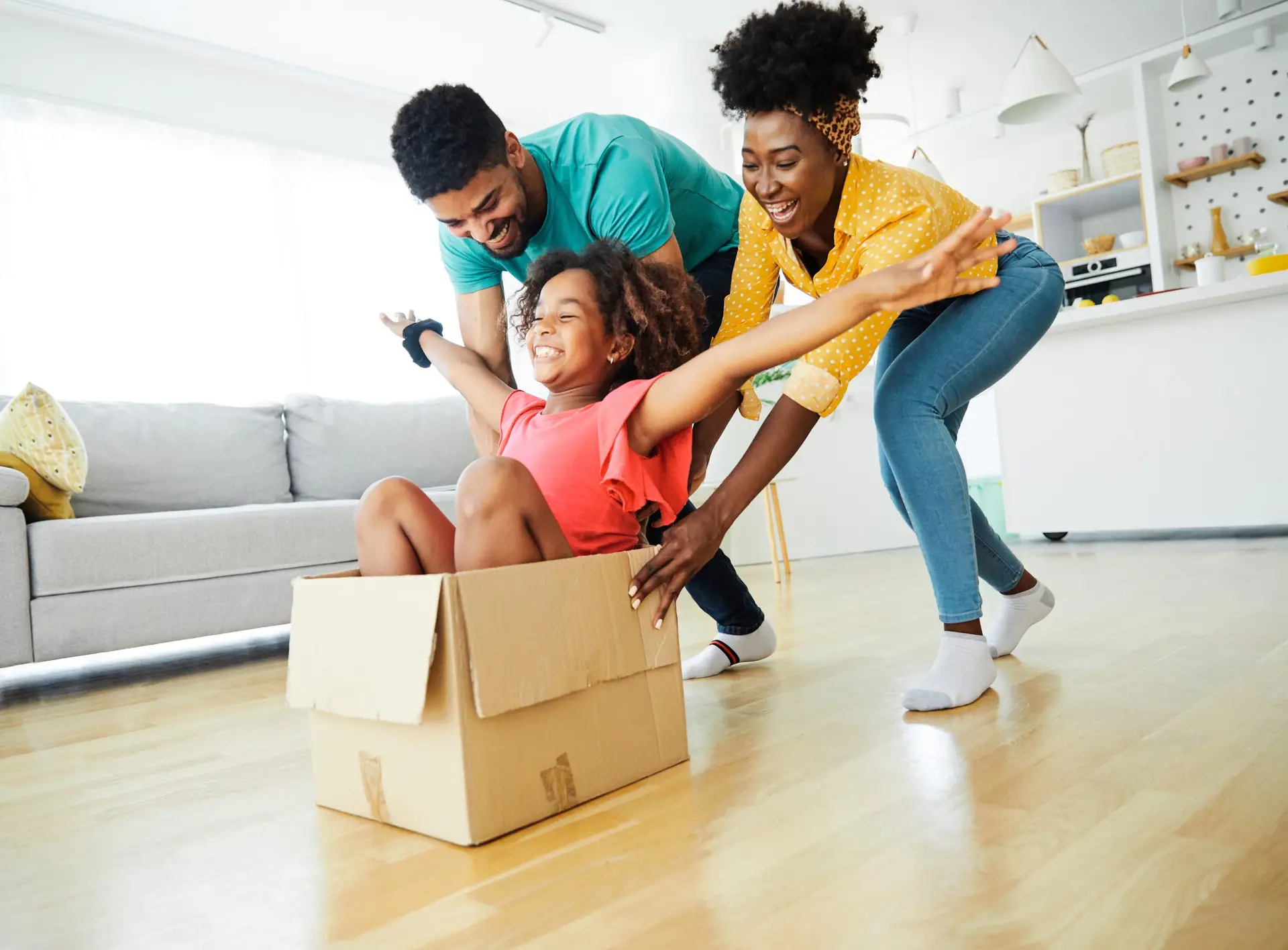 A joyful family in a bright living room enjoys some Family Fun. A child sits in a cardboard box with arms outstretched, pushed by two smiling adults, making weekend activities for families unforgettable. The scene conveys happiness and playful energy against a backdrop of a cozy sofa and kitchen area.