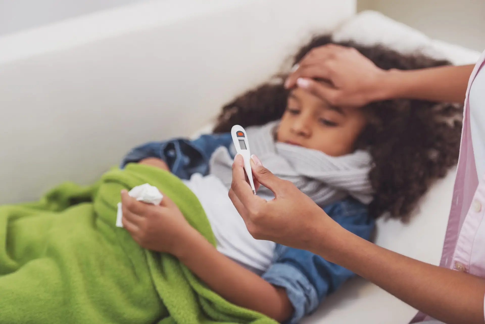 A child with curly hair, wrapped in a green blanket and wearing a scarf, lies on the couch. They're holding a tissue while an adult gently places a hand on their forehead and checks the thermometer, attentively taking care of the sick child and showing concern for a potential fever.