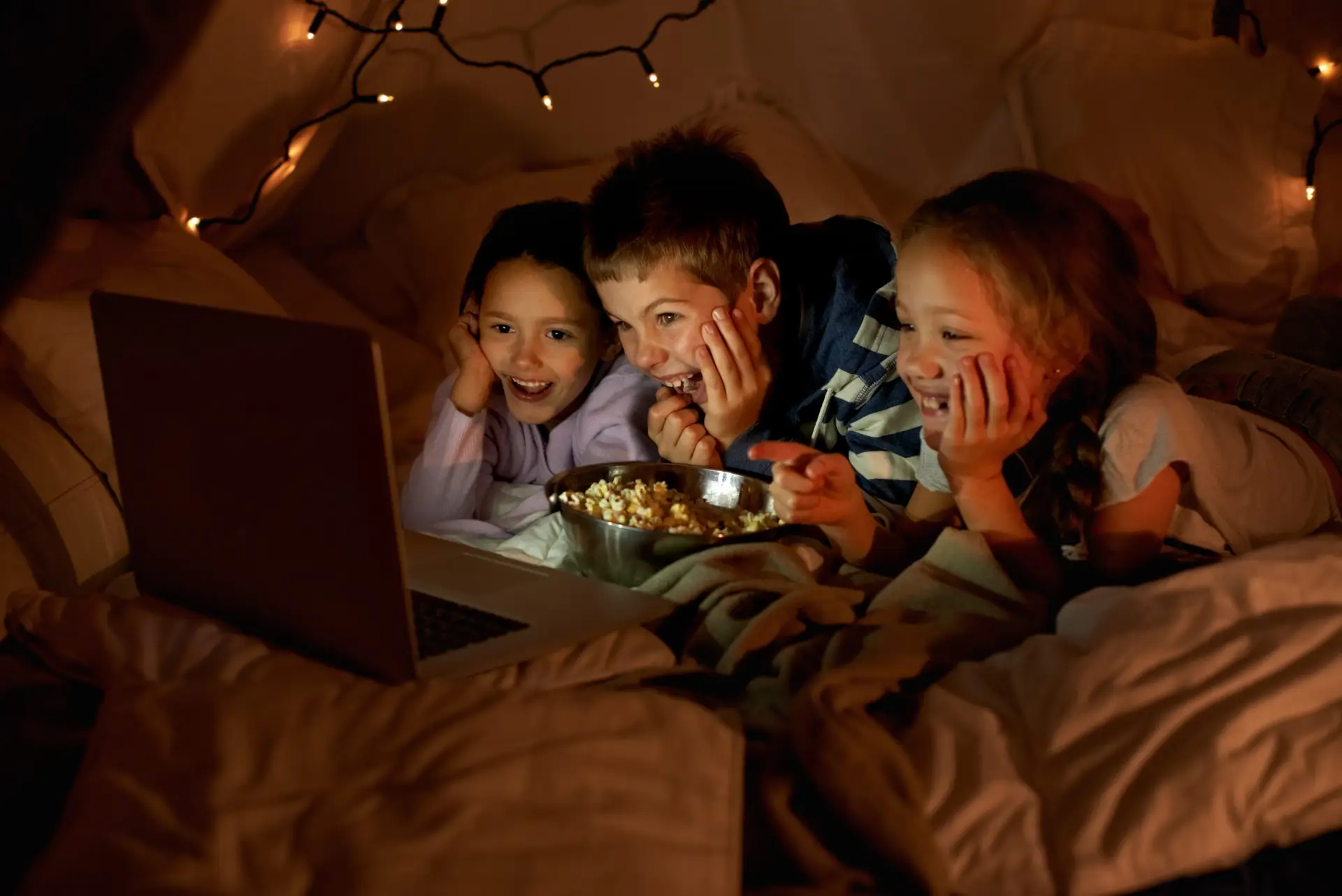 Three children are lying on a cozy bed, watching a laptop and smiling. Snuggled under blankets with a bowl of popcorn in front of them, they discuss scary sleepover games: harmful or harmless? Warm string lights in the background enhance the cozy atmosphere.